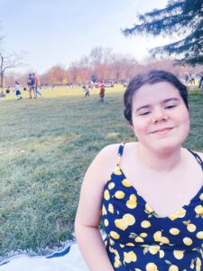 Lilian sits on the grass in a park, wearing a yellow and blue dress with a lemon pattern, while people enjoy the outdoors in the background.