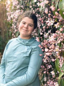 Lilian, in a light blue outfit, stands smiling against a backdrop of pink flowers and greenery. The scene conveys a cheerful and vibrant atmosphere.