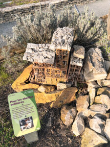 A structure made of stones and bricks designed as a habitat for solitary bees, surrounded by rocks and plants. A sign nearby provides information about its purpose.