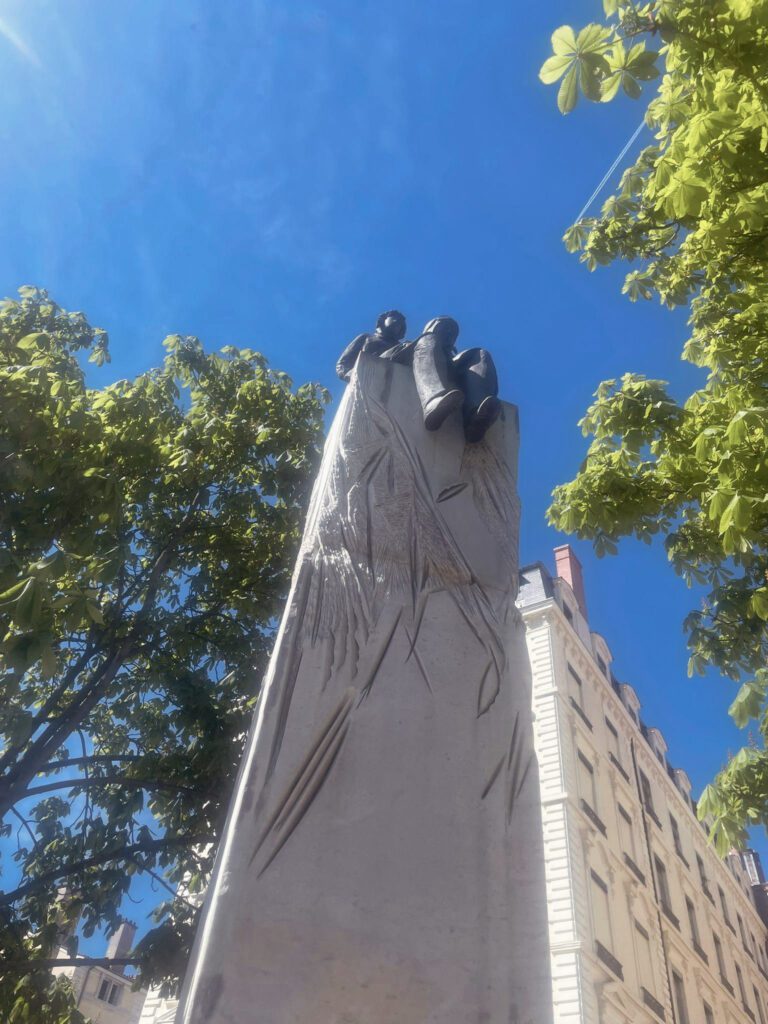 A tall monument with a sculpted hand reaching upward, surrounded by green trees and a clear blue sky. The base features intricate carvings, and a building is visible in the background.