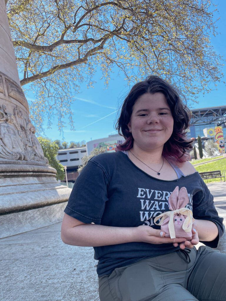 A person sits outdoors, smiling and holding a decorative bunny with pink ears. The background features trees and a clear blue sky.