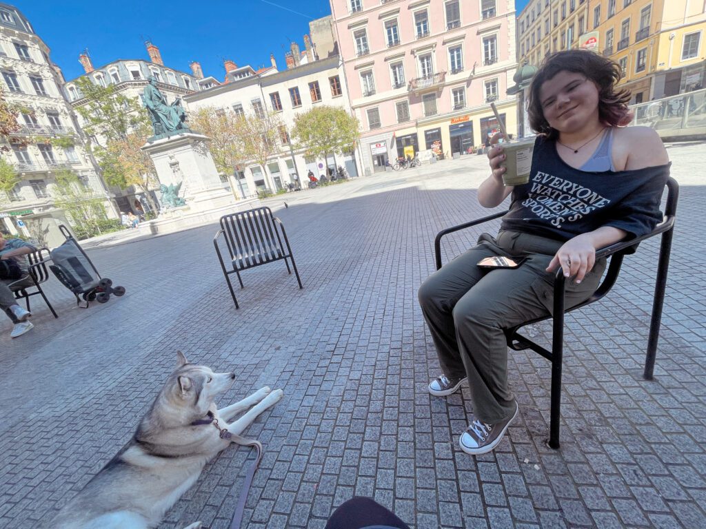 A woman sits in a chair with a cup in her hand, smiling at the camera, while a dog lies on the ground beside her in a sunny square surrounded by buildings.