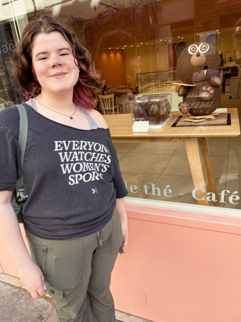 A young woman stands outside a café, smiling while wearing a shirt that reads "EVERYONE WATCHES WOMEN'S SPORT." Next to her is a large chocolate sculpture of a bear.