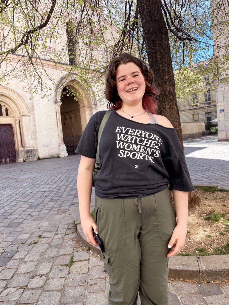 A young woman stands outdoors, smiling while wearing a black t-shirt that reads "EVERYONE WATCHES WOMEN'S SPORTS" and olive green pants. The background features a historic building and trees.