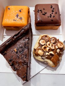Four assorted desserts are displayed in a box: a bright orange square topped with flowers, a glossy chocolate square, a triangular chocolate cake, and a round pastry topped with nuts and fruit.