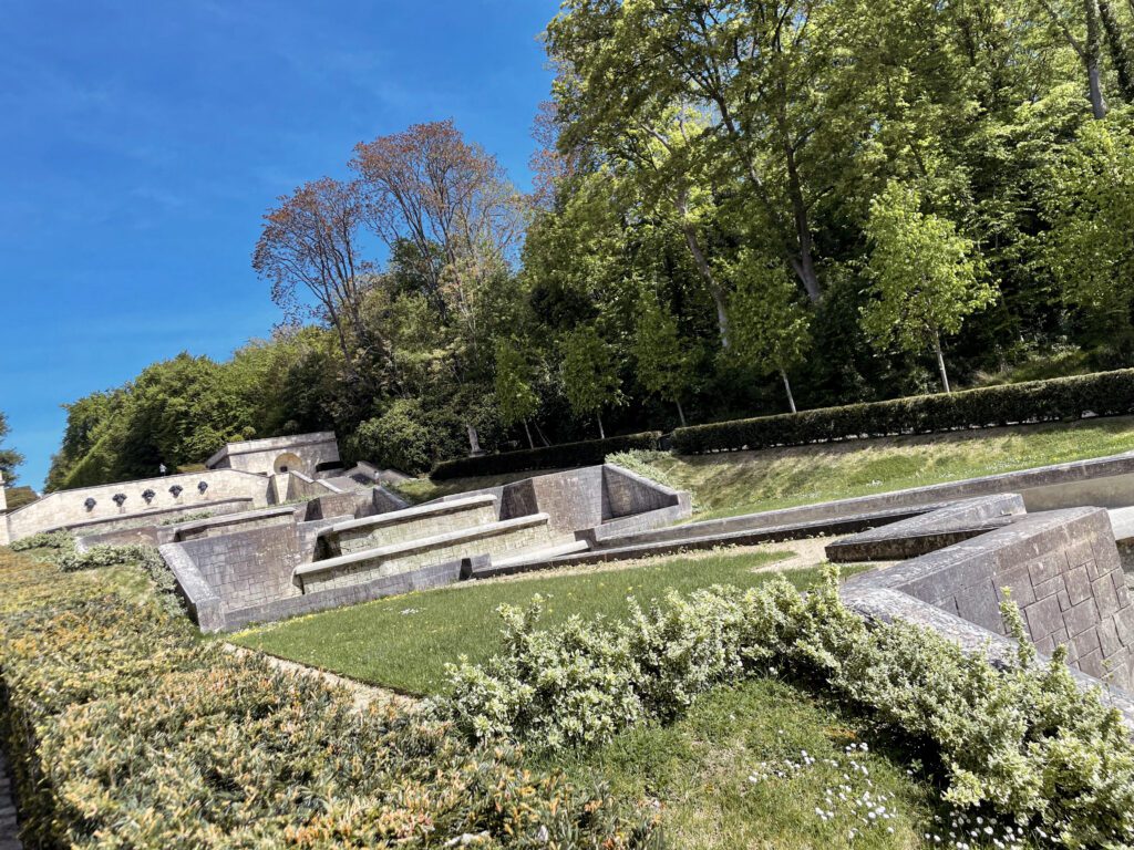 A landscaped garden with geometric stone pathways and green hedges, surrounded by trees under a clear blue sky.