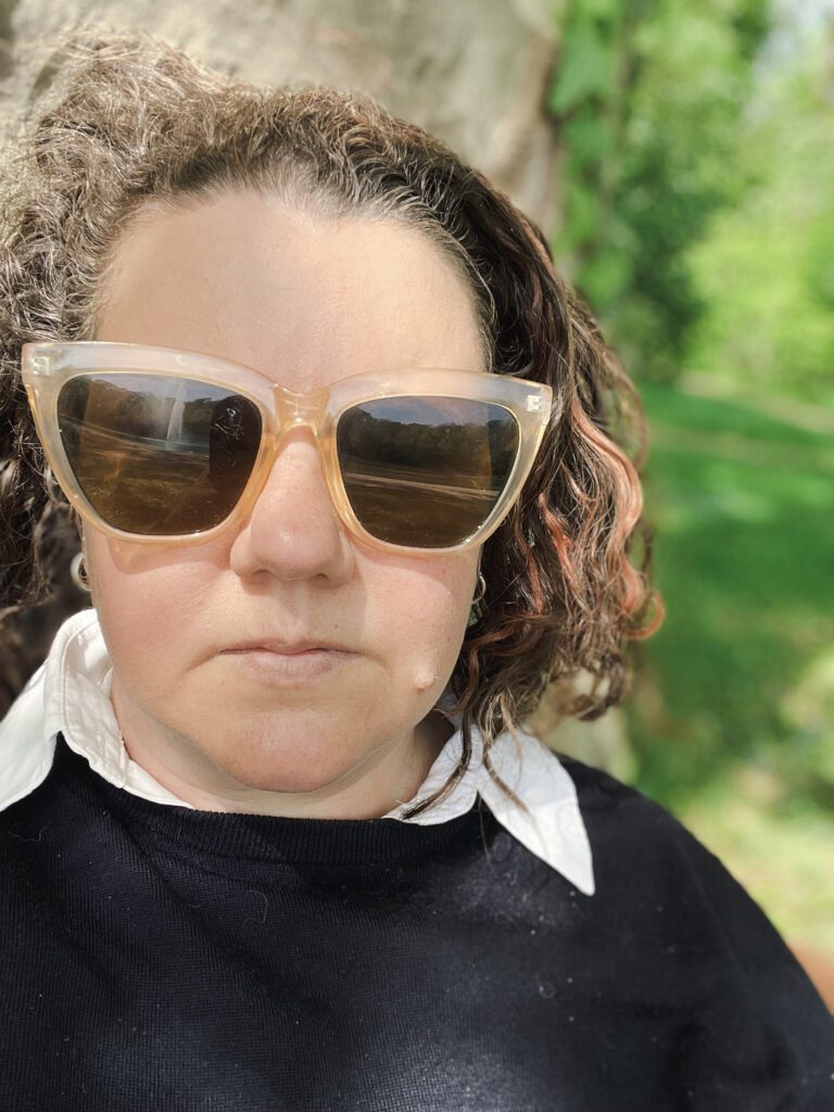 A person with curly hair (Hope) wears large, stylish sunglasses and a navy blue sweater, sitting in front of a blurred green background. The expression is serious, suggesting a moment of contemplation.