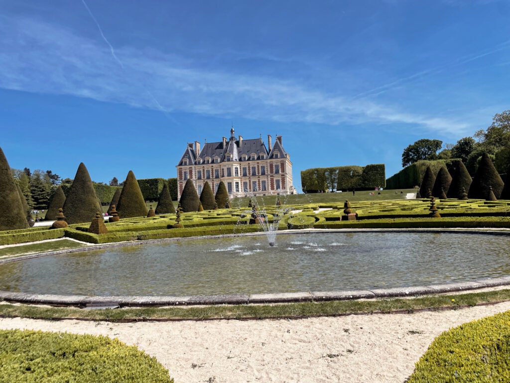 A grand chateau is surrounded by neatly trimmed hedges and a decorative garden, with a fountain in the foreground under a clear blue sky.