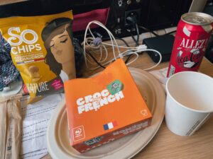 A tray with a box of croque monsieur, a bag of chips, a can of soda, and a paper cup, all set on a table with visible wires and documents in the background.