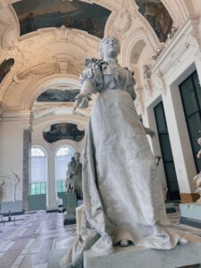 A detailed statue of a woman (Marie Deraismes) stands prominently in the Petit Palais, surrounded by ornate architecture and other sculptures. The bright interior highlights the artistic features of the museum.