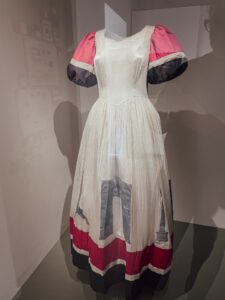 A white dress with red puffed sleeves and a red and blue hem displayed on a mannequin. The dress symbolizes celebration and liberation.