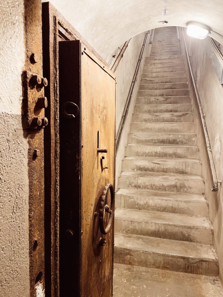 A metal fire door stands ajar, leading to a dimly lit stairway that ascends into a tunnel. The walls are bare and the stairs are worn.