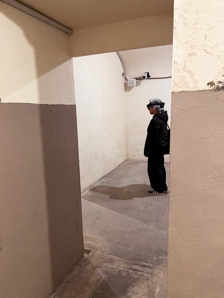 A person stands in a dimly lit corridor, wearing a headset, with walls painted in light and dark colors.