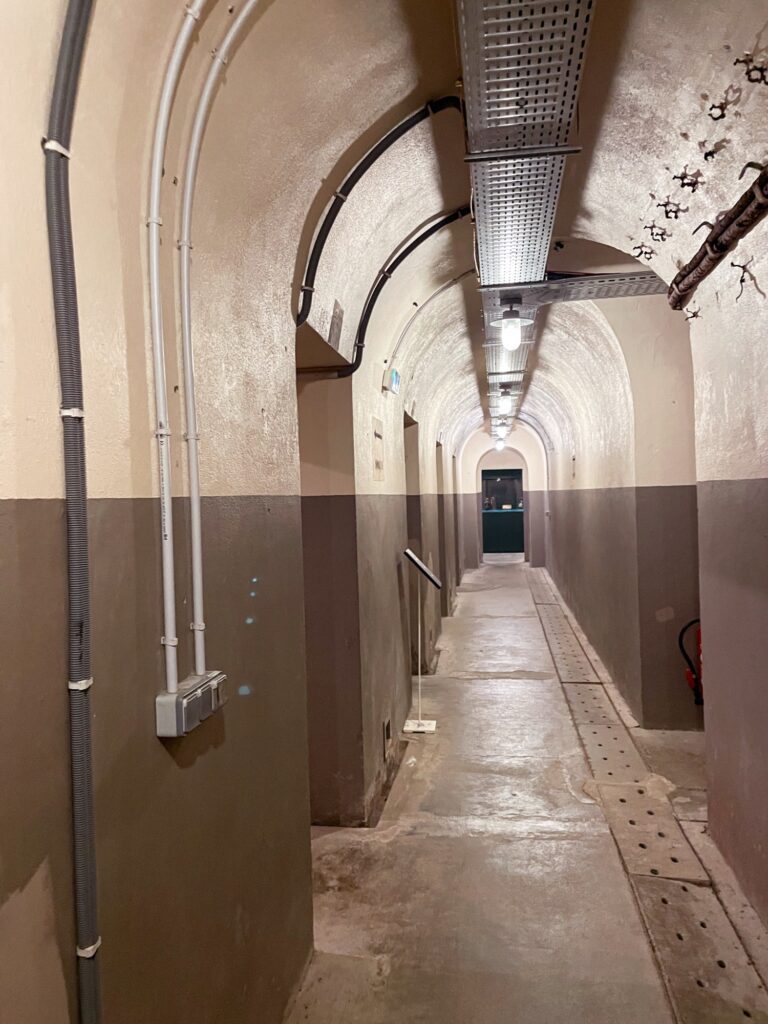 A long, narrow corridor with arched ceilings, featuring beige walls with a darker lower section. The floor is concrete, and there are pipes running along the ceiling, leading to a green door at the far end.