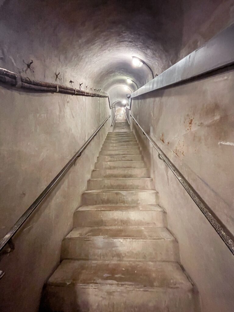 A dimly lit staircase leads upward through a narrow tunnel, with concrete walls and a railing on one side.