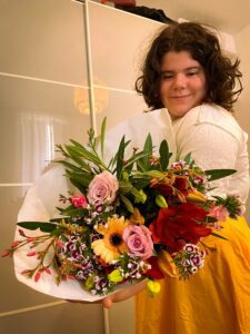 A young person (Lilian) smiles while holding a large bouquet of colorful flowers. The setting is indoors, with a light-colored background.