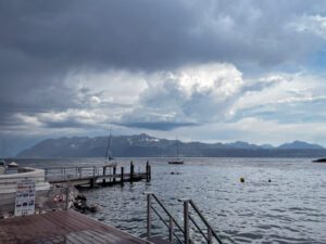 A scenic view of a lake with a wooden pier extending into the water, surrounded by mountains under a cloudy sky. The calm water reflects the clouds, creating a serene atmosphere.