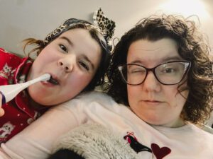 A woman and a girl sit closely together, both making playful expressions. The girl is holding a toothbrush, while the woman wears glasses and has curly hair.