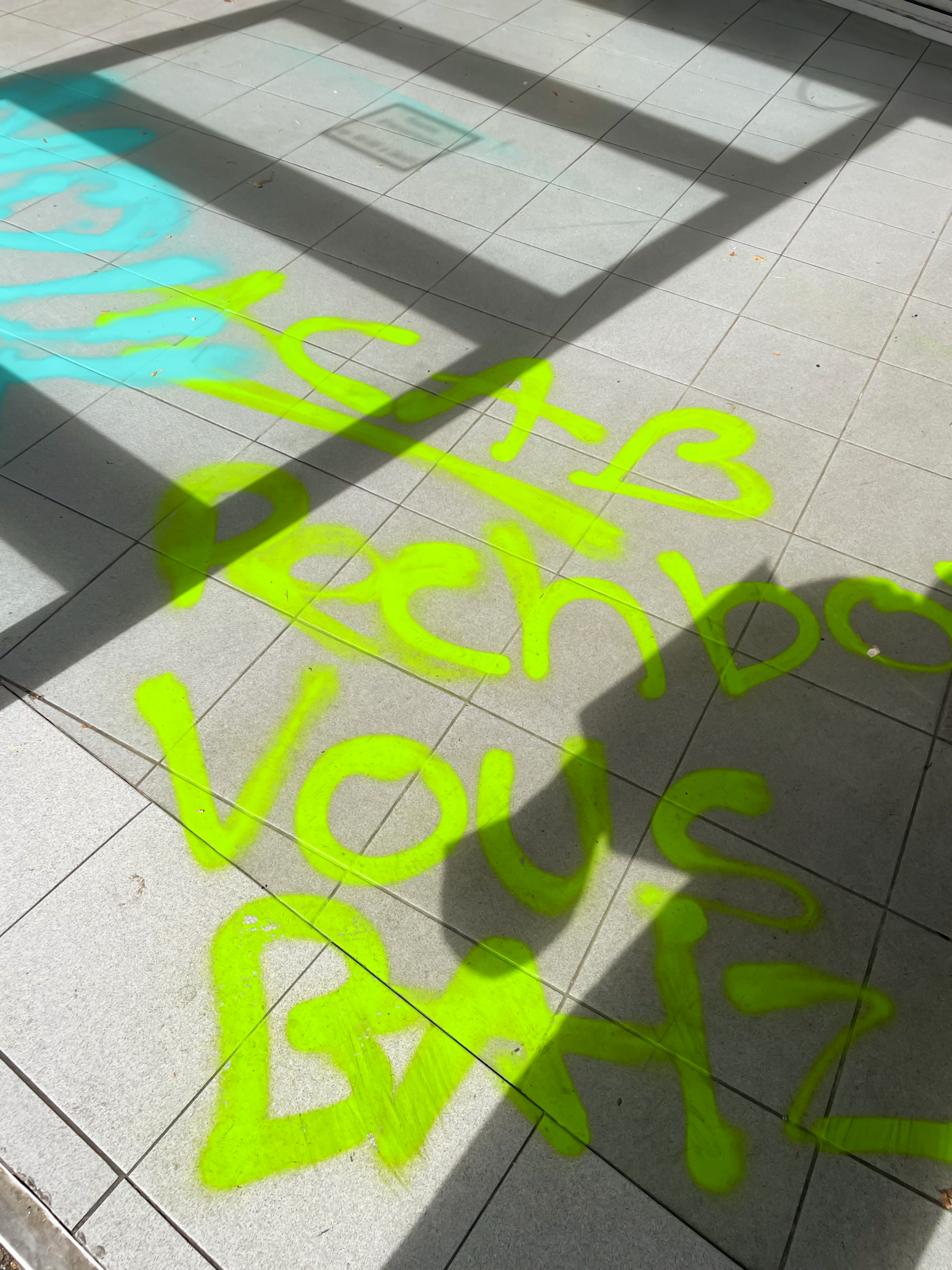 Bright green graffiti on a tiled floor with text that appears to convey a message related to student protests. The sunlight casts shadows on the tiles, highlighting the vibrant colors of the paint.