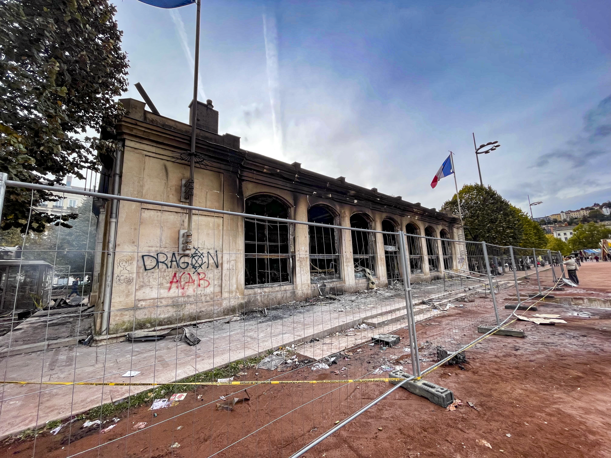 A stone building with graffiti and a French flag stands behind metal barriers on a muddy, debris-strewn street.
